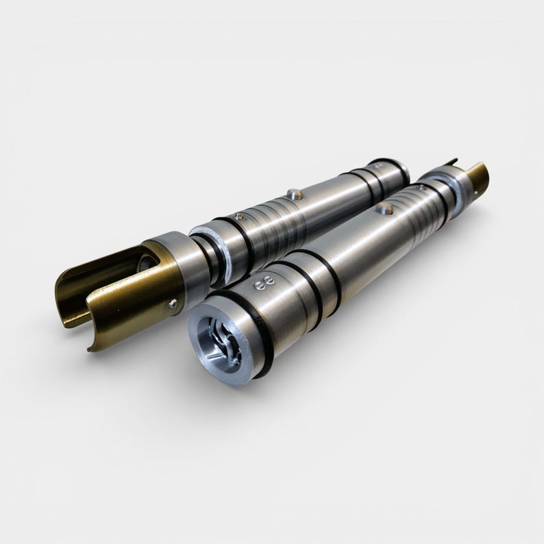 May include: Two metallic lightsaber hilts. One hilt features a gold-colored emitter and the other has a silver-colored emitter. Both hilts have a cylindrical design with black rings and silver accents. The hilts are lying on a white surface.