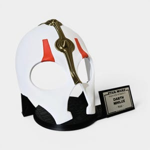 May include: A white Darth Nihilus mask with red accents and gold detailing. The mask is displayed on a black stand with a Star Wars plaque that reads "Darth Nihilus - Mask".