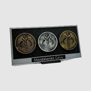 May include: Three Mandalorian coins displayed in a black frame with the text "Mandalorian Coins". The coins are silver, gold, and bronze and feature a stylized image of a Mandalorian symbol.