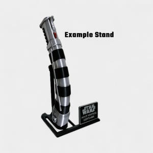 May include: A silver and black lightsaber with a red button, displayed on a black stand. The stand has a small plaque with "STAR WARS" and "ASAJI VENTRESS LIGHTSABER" printed on it. The text "Example Stand" is also visible.