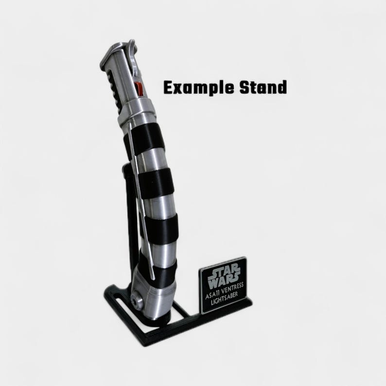 May include: A silver and black lightsaber with a red button, displayed on a black stand. The stand has a small plaque with "STAR WARS" and "ASAJI VENTRESS LIGHTSABER" printed on it. The text "Example Stand" is also visible.