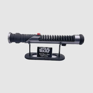 May include: A black and silver Star Wars lightsaber replica with a red button on the handle. The lightsaber is on a black stand with a silver plaque that reads "Star Wars Qui-Gon Jinn Lightsaber".