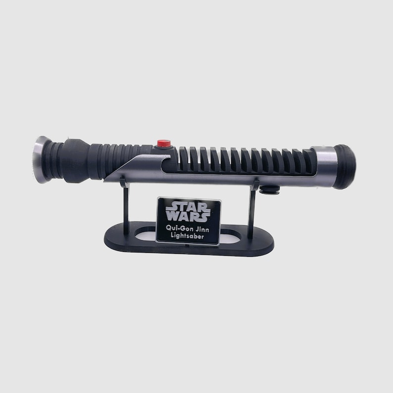May include: A black and silver Star Wars lightsaber replica with a red button on the handle. The lightsaber is on a black stand with a silver plaque that reads "Star Wars Qui-Gon Jinn Lightsaber".