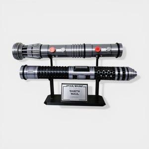 Puede incluir: Dos sables de luz sobre un soporte negro. El sable de luz superior es plateado con botones rojos. El sable de luz inferior es negro y plateado. Una placa dice "Star Wars Darth Maul Lightsaber".