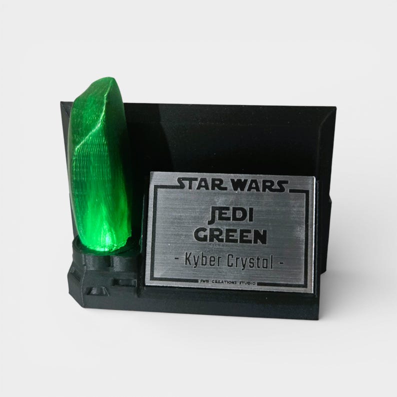 May include: A black display stand featuring a glowing green Kyber Crystal. A silver plaque reads "STAR WARS JEDI GREEN - Kyber Crystal." The stand is designed to hold the crystal and is a collectible item.