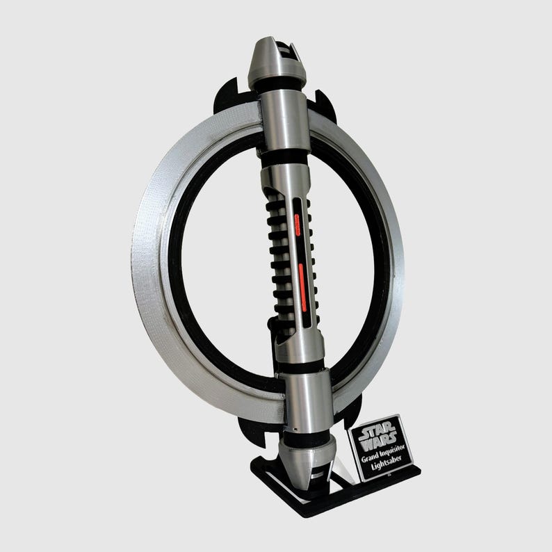 Puede incluir: Un sable de luz plateado y negro con una hoja roja, exhibido en un soporte con una base circular. El soporte tiene una placa plateada con el texto "Star Wars Grand Inquisitor Lightsaber".