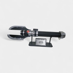 May include: A black and silver Darth Malgus lightsaber replica, displayed on a black stand. The lightsaber features a red button and a unique blade design. The stand includes a Star Wars plaque.