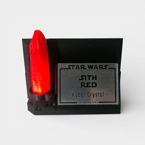 May include: A black display stand featuring a glowing red Kyber Crystal, labeled "Sith Red" from Star Wars. The stand has a metallic nameplate with the Star Wars logo and text.