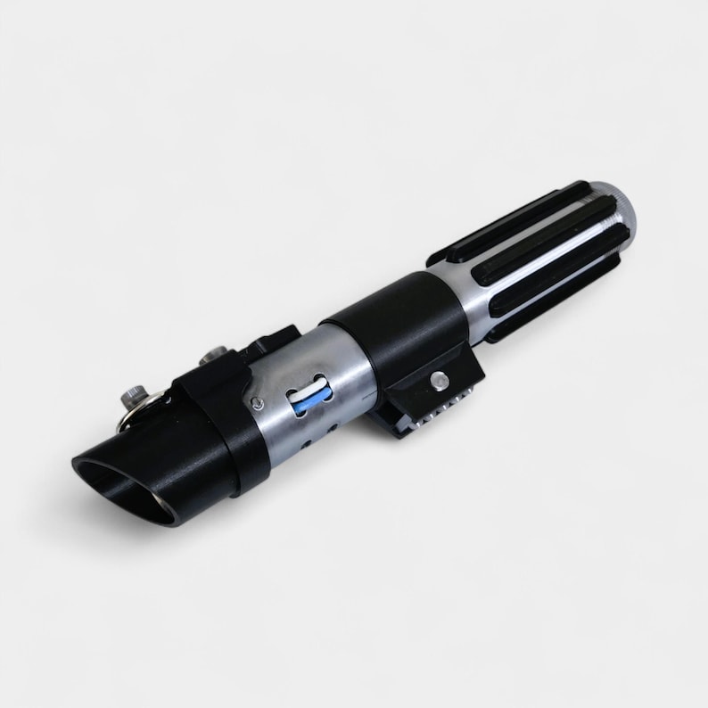 May include: A black and silver lightsaber prop with a cylindrical design. The hilt features a black grip, silver accents, and a ribbed end. A blue light is visible near the base.
