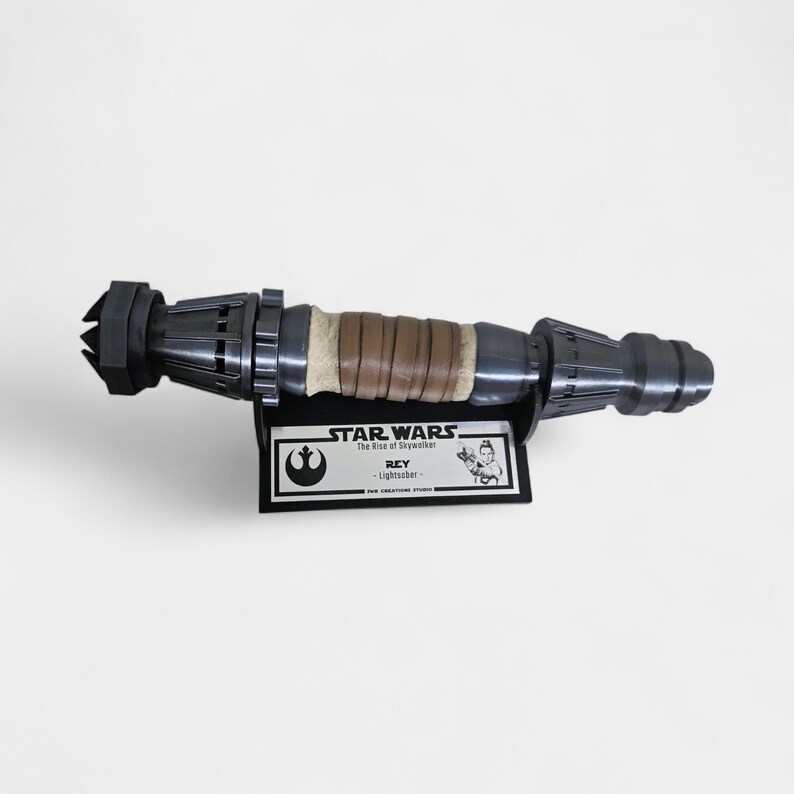 May include: A gray lightsaber prop from Star Wars: The Rise of Skywalker, with a brown leather wrap and a black display stand. The stand has a label that reads "STAR WARS The Rise of Skywalker REY - Lightsaber -", and a small image of Rey holding the lightsaber.