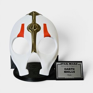May include: A white Darth Nihilus mask with gold and red detailing, presented on a black stand. The mask features a central gold design and red side elements. A silver plaque with "STAR WARS Darth Nihilus Mask" is beside the mask.