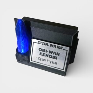 May include: A black display stand featuring a blue Kyber Crystal. The stand has a silver plaque with the text "STAR WARS OBI-WAN KENOBI - Kyber Crystal". The display is designed for Star Wars fans.