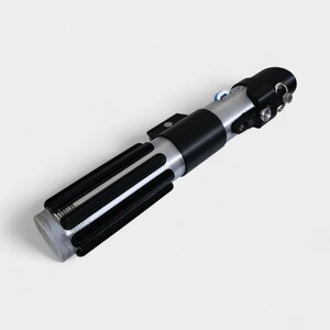 May include: A replica lightsaber with a black and silver handle. The hilt features a ribbed grip, a silver midsection, and a black emitter. The design includes various buttons and details, resembling a prop from a science fiction film.