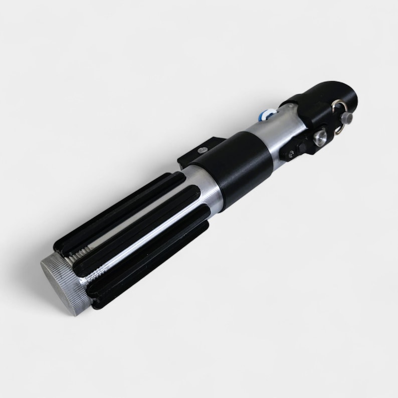 May include: A replica lightsaber with a black and silver handle. The hilt features a ribbed grip, a silver midsection, and a black emitter. The design includes various buttons and details, resembling a prop from a science fiction film.