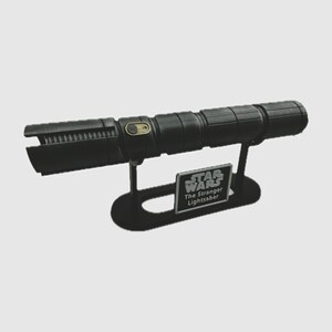 May include: A black lightsaber with a gold button on the handle, displayed on a black stand with a silver plaque that reads "Star Wars The Stranger Lightsaber".