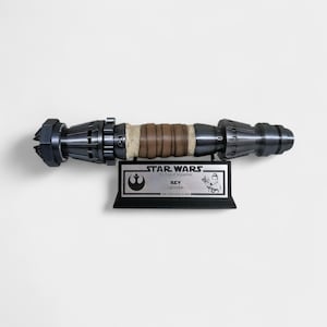 May include: A black and silver Star Wars lightsaber prop replica with a brown leather-wrapped handle. The prop is mounted on a black plaque with a silver Star Wars logo and the text "STAR WARS The Rise of Skywalker REY Lightsaber FOR TREKSTORE STUDIO".