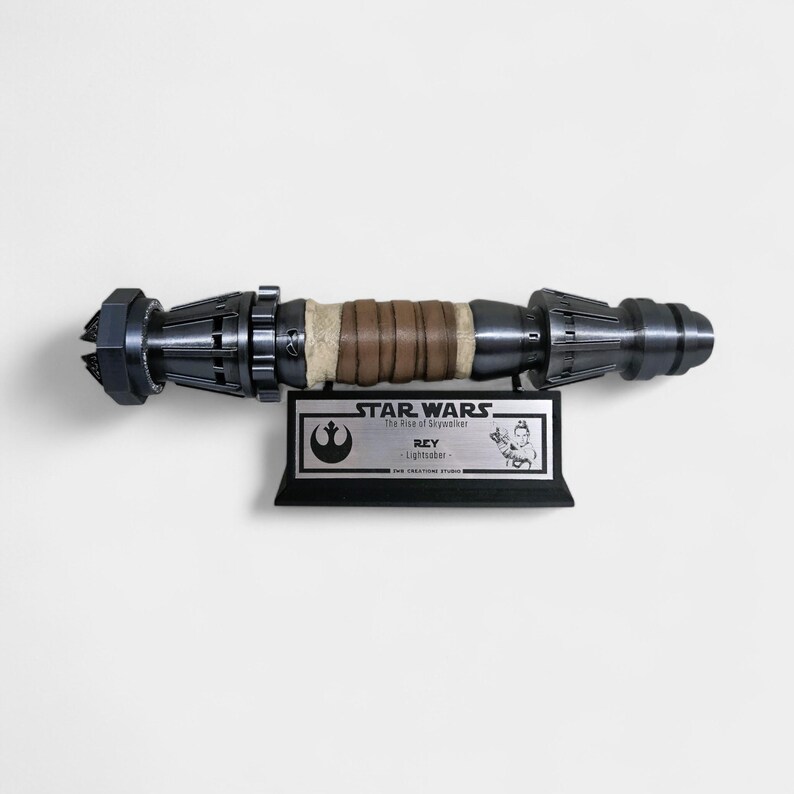 May include: A black and silver Star Wars lightsaber prop replica with a brown leather-wrapped handle. The prop is mounted on a black plaque with a silver Star Wars logo and the text "STAR WARS The Rise of Skywalker REY Lightsaber FOR TREKSTORE STUDIO".