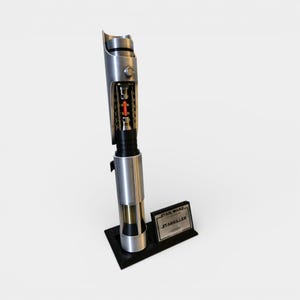 May include: A silver and black Star Wars Starkiller lightsaber replica. The lightsaber is displayed on a black stand with a nameplate that reads "STAR WARS STARKILLER Lightsaber". The lightsaber has a detailed internal mechanism with red and gold accents.