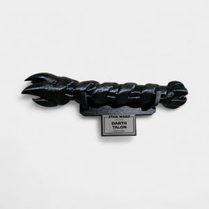 May include: A black Darth Talon lightsaber from Star Wars Legends, mounted on a black display stand. The lightsaber has a textured, detailed design with a claw-like end. The display stand features the Star Wars logo and the name "Darth Talon".