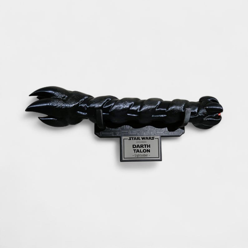 May include: A black Darth Talon lightsaber from Star Wars Legends, mounted on a black display stand. The lightsaber has a textured, detailed design with a claw-like end. The display stand features the Star Wars logo and the name "Darth Talon".