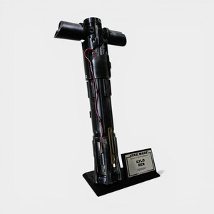 May include: A black Kylo Ren lightsaber replica, featuring a crossguard design. The prop is displayed on a black stand with a nameplate that reads "STAR WARS KYLO REN - Lightsaber". Red wires are visible, adding detail to the prop.