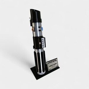 May include: A replica of Darth Vader's lightsaber, featuring a black handle and silver accents. The lightsaber is mounted on a black base with a small plaque that reads "STAR WARS Darth Vader Lightsaber". The lightsaber is a prop from the Star Wars franchise.
