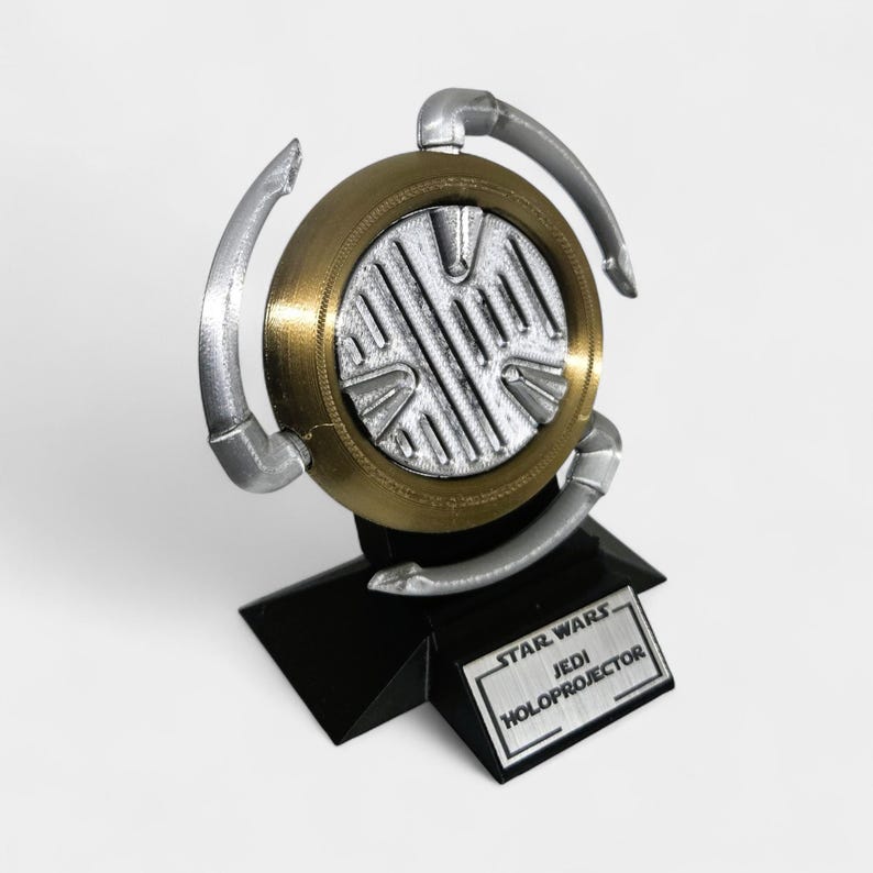 Op de afbeelding: Een goud-zilveren Jedi Holoprojector model uit Star Wars. Het model heeft een cirkelvormig ontwerp met zilveren accenten en een zwarte basis. De tekst "STAR WARS JEDI HOLOPROJECTOR" staat op een kleine rechthoekige plaat.
