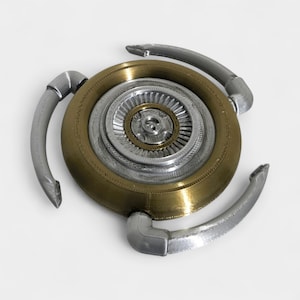 Op de afbeelding: Een metalen, drietandig object met een centraal cirkelvormig ontwerp. Het midden heeft concentrische cirkels in zilver- en goudtinten, met drie gebogen zilveren armen die naar buiten uitsteken. Het object heeft een mechanische of industri&euml;le uitstraling.