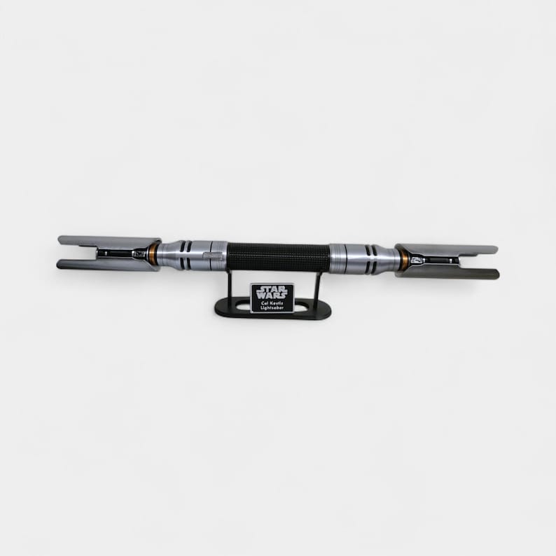 May include: A silver and black lightsaber with a black handle, displayed on a black stand. The lightsaber has a textured grip and silver accents. The stand has a Star Wars logo and text that reads "Cal Kestis Lightsaber."