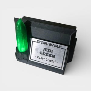 May include: A black display stand featuring a vibrant green Kyber Crystal. The stand has a silver plaque with the words "STAR WARS JEDI GREEN - Kyber Crystal". The display is designed to showcase the crystal.