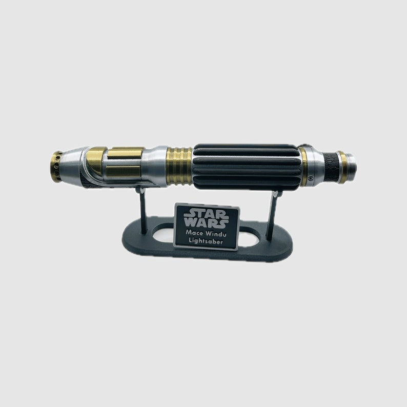 May include: A black, silver and gold lightsaber replica on a black stand with a silver plaque that reads "STAR WARS Mace Windu Lightsaber".