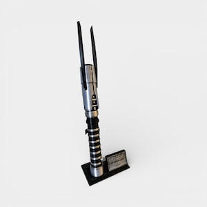 Op de afbeelding: Een zilver-zwarte lichtzwaard replica met twee zwarte bladen die zich naar boven uitstrekken. Het handvat heeft zilveren en zwarte details, met een zwarte basis en een klein plaatje met de tekst "STAR WARS STARKILLER".