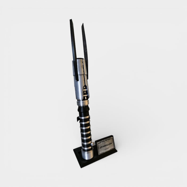 Op de afbeelding: Een zilver-zwarte lichtzwaard replica met twee zwarte bladen die zich naar boven uitstrekken. Het handvat heeft zilveren en zwarte details, met een zwarte basis en een klein plaatje met de tekst "STAR WARS STARKILLER".