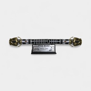 May include: A black lightsaber with gold accents and a silver blade. The lightsaber is on a black display stand with a silver plaque that reads "STAR WARS: JEDI SURVIVOR - DAGAN GERA - LIGHTSABER".