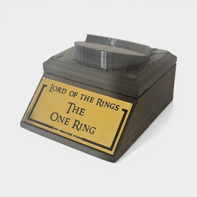 May include: A dark gray box with a gold plaque that reads "Lord of the Rings The One Ring." The top of the box has a curved, gray design. The box is a decorative item.