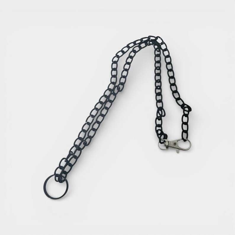 May include: Black chain lanyard with a silver-colored clasp and a black ring. The chain is made of small, interconnected links. The lanyard is designed to hold keys or other small items.