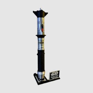 May include: A silver and black lightsaber with a black stand and a silver plaque that reads "Star Wars Baylan Skoll Lightsaber".