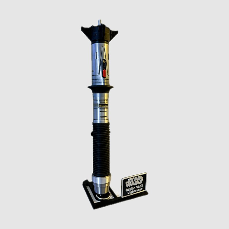 May include: A silver and black lightsaber with a black stand and a silver plaque that reads "Star Wars Baylan Skoll Lightsaber".