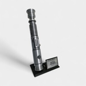May include: A silver lightsaber prop with a black base and a small plaque that reads "STAR WARS, AAYLA SECURA, Lightsaber". The lightsaber has a detailed design with a ribbed handle and a button.
