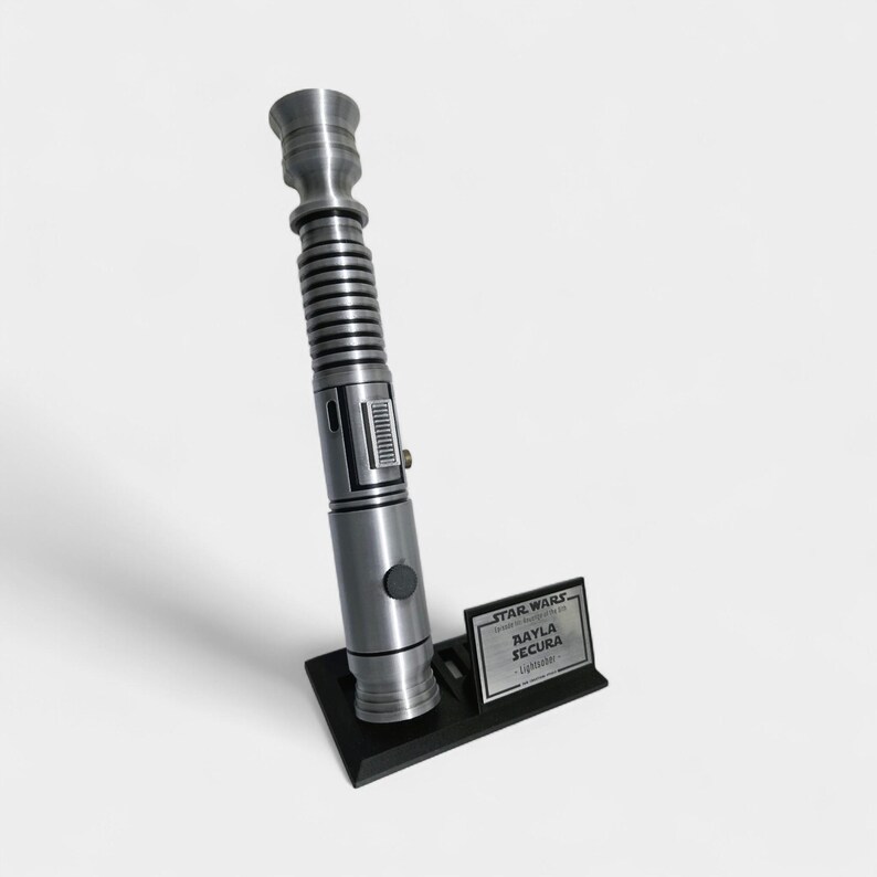 May include: A silver lightsaber prop with a black base and a small plaque that reads "STAR WARS, AAYLA SECURA, Lightsaber". The lightsaber has a detailed design with a ribbed handle and a button.