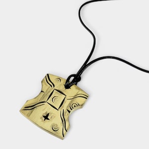 May include: A gold-coloured pendant with a black cord. The pendant features a square and star design with curved lines and circular patterns. The pendant is made of a light-coloured material.
