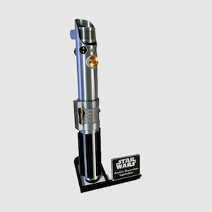 May include: A silver-toned metal lightsaber replica with black vertical grip panels, a gold-colored circular activation button, and a textured mid-section. It stands on a black display base featuring a plaque that reads STAR WARS Anakin Skywalker Lightsaber.