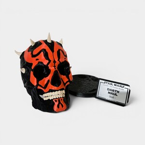 Op de afbeelding: Een zwart en rood Darth Maul schedelbeeldje met witte tanden en hoorns. Het beeldje staat naast een zwarte basis en een wit label met de tekst "STAR WARS Darth Maul - Skull".