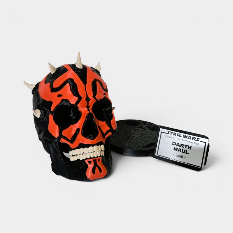 Op de afbeelding: Een zwart en rood Darth Maul schedelbeeldje met witte tanden en hoorns. Het beeldje staat naast een zwarte basis en een wit label met de tekst "STAR WARS Darth Maul - Skull".