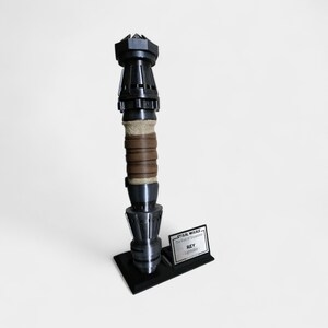 May include: A black and silver lightsaber replica with a brown leather wrap on the handle. The lightsaber is on a black stand with a silver plaque that reads "STAR WARS The Rise of Skywalker  REY Lightsaber".