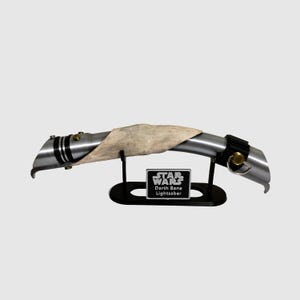 May include: A silver lightsaber with a black handle and a white leather wrap. The lightsaber is on a black stand with a silver plaque that reads "STAR WARS Darth Bane Lightsaber".