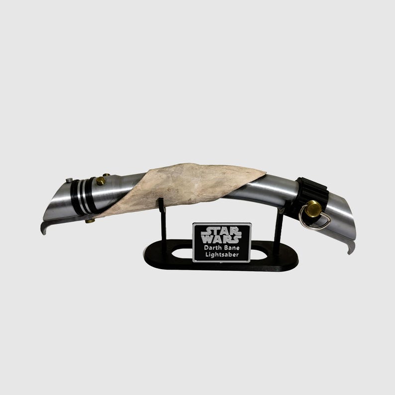 May include: A silver lightsaber with a black handle and a white leather wrap. The lightsaber is on a black stand with a silver plaque that reads "STAR WARS Darth Bane Lightsaber".