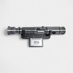 May include: A silver lightsaber prop, mounted on a black display stand. The stand features a nameplate that reads "Aayla Secura - Lightsaber" with the Star Wars logo. The lightsaber has a detailed, metallic design.