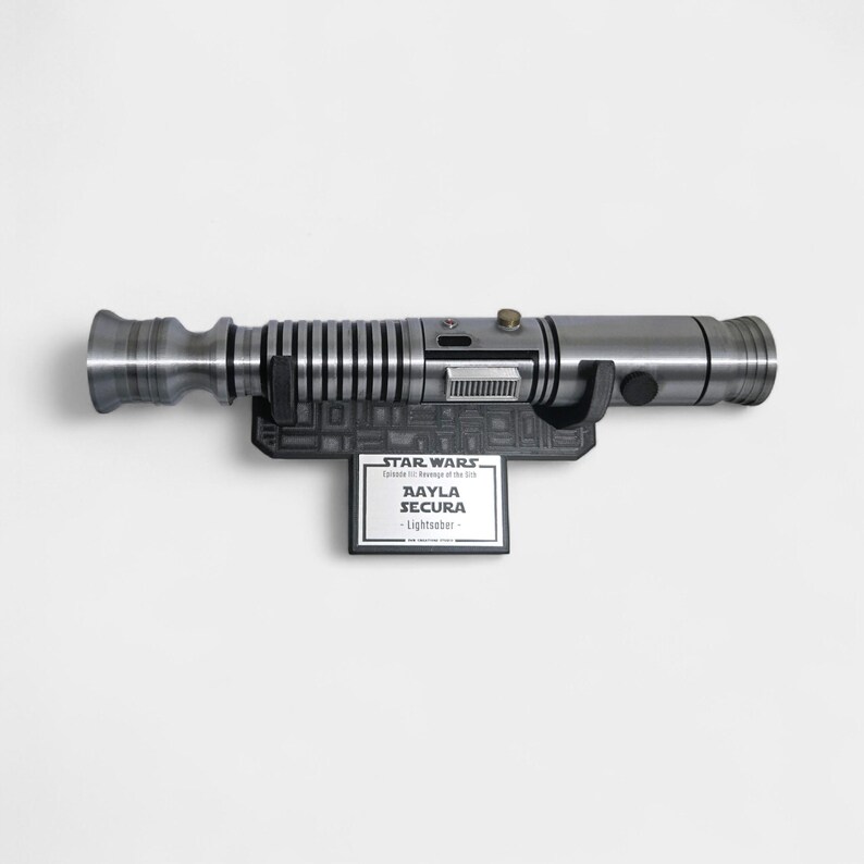 May include: A silver lightsaber prop, mounted on a black display stand. The stand features a nameplate that reads "Aayla Secura - Lightsaber" with the Star Wars logo. The lightsaber has a detailed, metallic design.