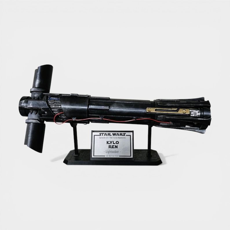 May include: A black Kylo Ren lightsaber prop from Star Wars, Episode VII: The Force Awakens. The lightsaber has a unique crossguard design and is displayed on a black stand with a nameplate that reads "Kylo Ren - Lightsaber".